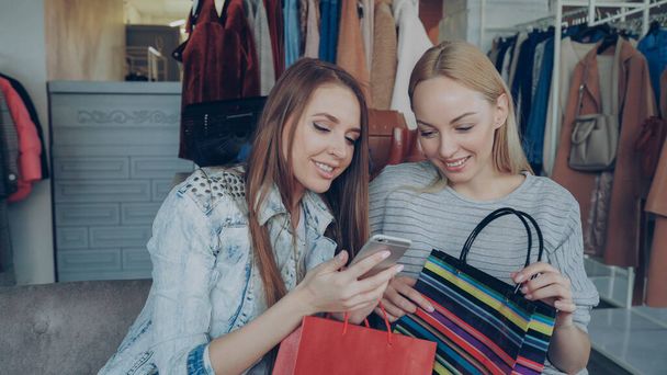 Attractive young ladies sitting on chairs in a clothing store with coffee and shopping bags, checking smartphone and talking. Shelves and hangers with colourful clothes in background - Foto, imagen