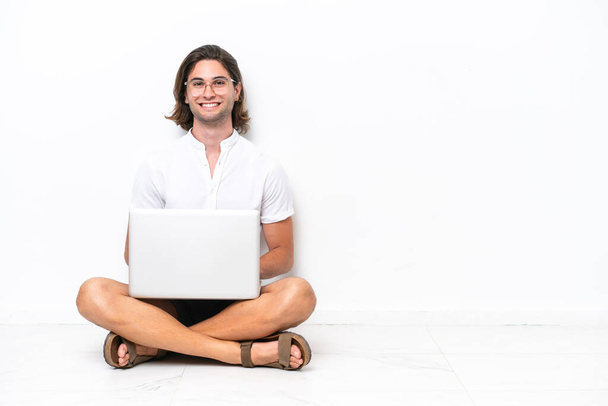Yerde dizüstü bilgisayarı olan yakışıklı genç bir adam beyaz arka planda izole bir şekilde oturuyor ve kollarını ön cephede çapraz tutuyor. - Fotoğraf, Görsel