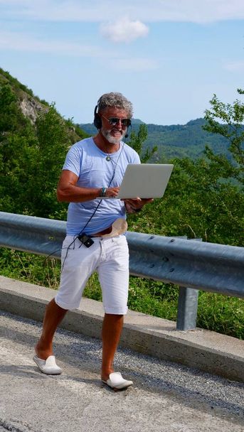 Gray-haired elderly man diabetic sick freelancer with a beard in sunglasses, working on a laptop on nature, mountains. A crazy and extraordinary old man walking to road in the summer. Vertical - Foto, imagen