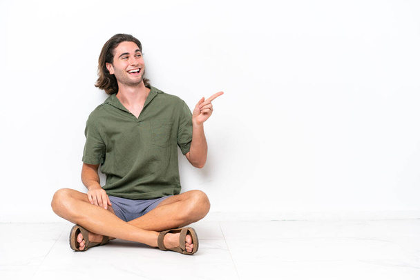 Mladý pohledný muž sedící na podlaze izolovaný na bílém pozadí ukazující prstem na stranu a prezentující produkt - Fotografie, Obrázek