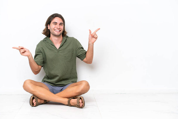 Mladý pohledný muž sedí na podlaze izolované na bílém pozadí ukazuje prstem na laterály a šťastný - Fotografie, Obrázek
