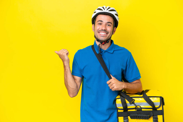 Молодий красивий чоловік з термальним рюкзаком над ізольованим жовтим тлом, що вказує на бік, щоб представити продукт
 - Фото, зображення