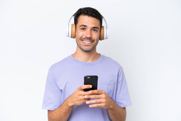 Молодий красивий чоловік над ізольованим білим тлом слухає музику з мобільним і дивиться спереду
 - Фото, зображення