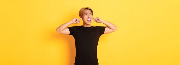 Portrait of annoyed and bothered asian guy grimacing, shut ears disturbed by loud noise, standing yellow background. - Photo, Image