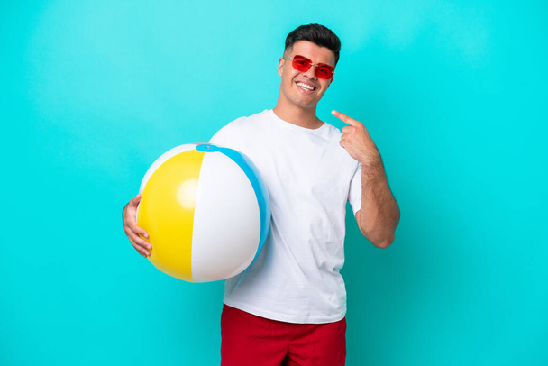 Mladý běloch drží plážový míč izolované na modrém pozadí dává palce nahoru gesto - Fotografie, Obrázek