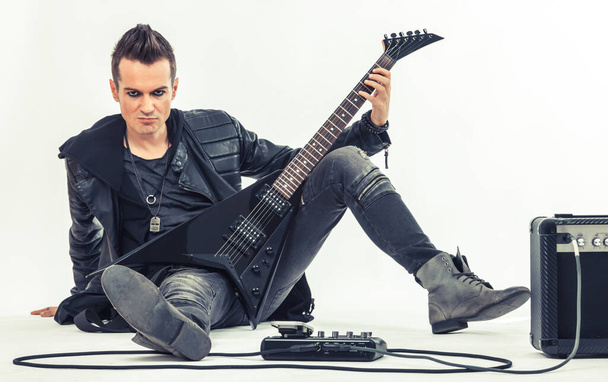 Музыкант хэви-метал сидит на полу, держа в руках электрогитару и глядя в камеру. - Фото, изображение