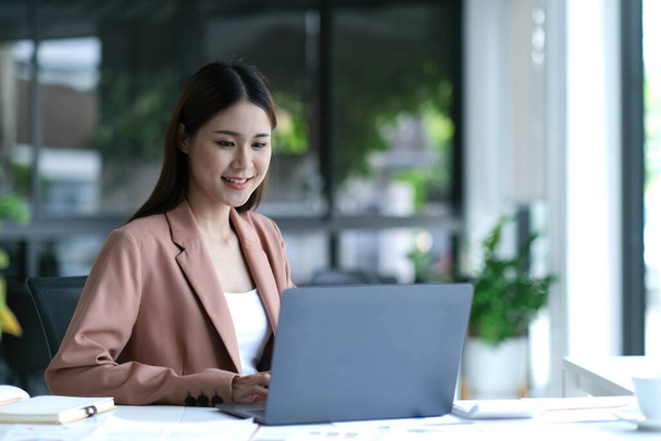 очаровательная азиатская деловая женщина, сидящая на ноутбуке в офисе - Фото, изображение