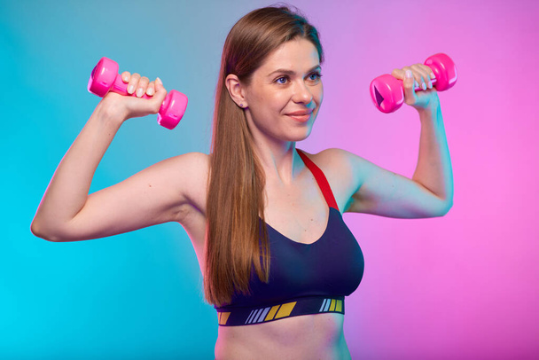smiling sporty woman in sports bra doing exercise with pink dumbbells looking away. Female fitness portrait isolated on neon multicolor background. - Φωτογραφία, εικόνα