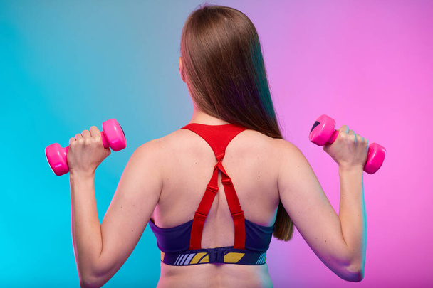 Sporty woman with long hair in fitness sportswear doing exercise with pink dumbbells back view. Female fitness portrait isolated on neon multicolor background. - Фото, зображення
