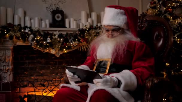 Moderni hymyilevä Santa Claus gamer pelaa mobiilipeliä tabletin taustalla takka, joulukuusi. Vanhus isoisä pukeutunut Joulupukki puku käyttää tabletti istuu tuolissa. Uudenvuoden vapaapäivät - Materiaali, video