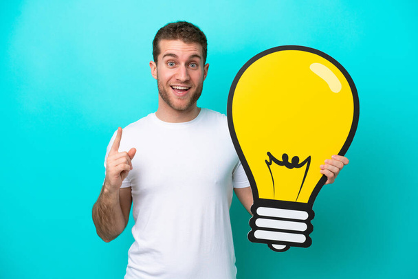 Young caucasian man isolated on blue background holding a bulb icon and thinking - Photo, Image