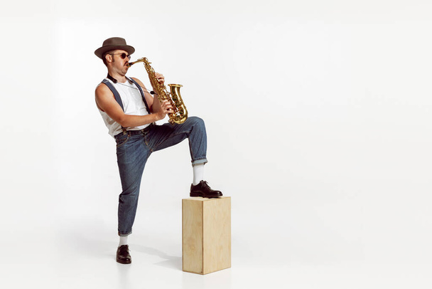 Portrait of stylish man in hat and suglasses playing saxophone , performing isolated over white background. Concept of live music, performance, retro style, creativity, artistic lifestyle, fashion - 写真・画像