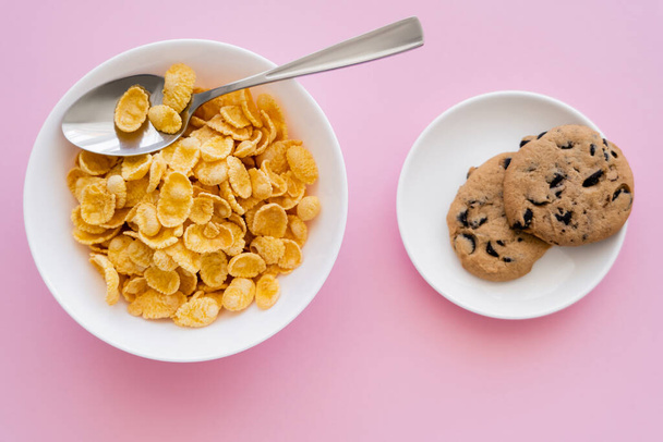 top view of bowl with tasty corn flakes and spoon near saucer with chocolate chip cookies on pink - 写真・画像