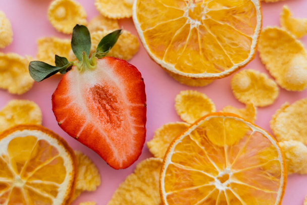 vista de cerca de fresa fresca en rodajas alrededor de copos de maíz crujientes y naranjas secas en rosa - Foto, Imagen