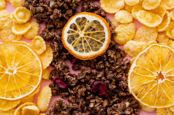 сверху вид хрустящих кукурузных хлопьев и вкусной мюсли рядом с сушеными апельсинами на розовом  - Фото, изображение
