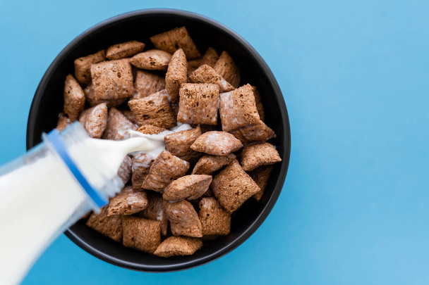 top view of milk pouring from bottle into bowl with cereal puffs isolated on blue - Foto, Imagen