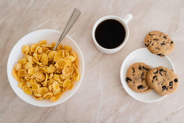 top view of bowl with corn flakes near cup of coffee and chocolate chip cookies on marble surface - Photo, Image