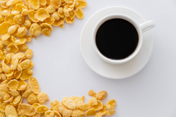 bovenaanzicht van zoete en krokante cornflakes in de buurt van een kopje zwarte koffie geïsoleerd op wit  - Foto, afbeelding