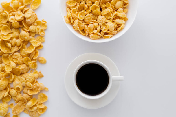 верхний вид чаши с кукурузными хлопьями возле чашки черного кофе изолированы на белом  - Фото, изображение