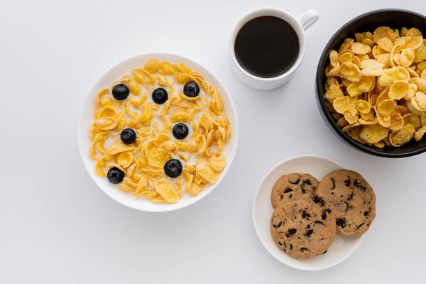 top view of bowls with corn flakes in milk with blueberries near cup of coffee and cookies isolated on white  - Photo, Image