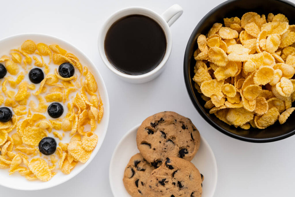 top view of bowls with corn flakes in milk with blueberries near cup of coffee and chocolate chip cookies isolated on white  - Foto, Bild