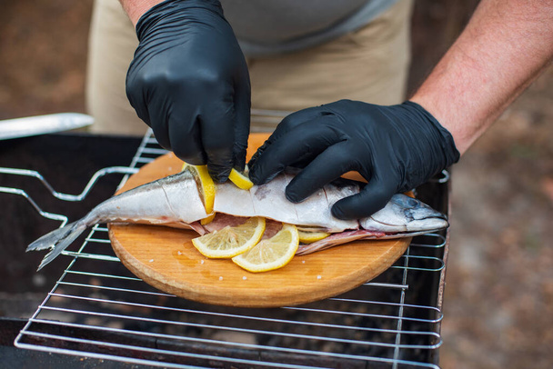 Grilled mackerel fish with lemon slices, top view. The process of cooking fish outdoors. Chef in black latex gloves preparing mackerel fish on a wooden cutting board. Tasty and fresh food, picnic, party, outdoor recreation - Foto, Bild
