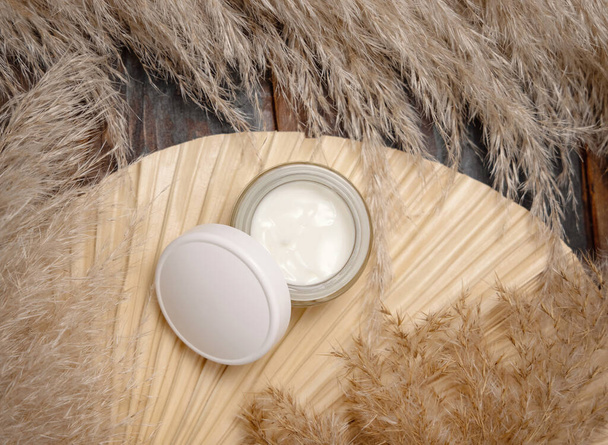 Opened cream jar with a blank lid on dried palm leaf near pampas grass top view, mockup. Skincare beauty product. Bohemian eco friendly cosmetics flat lay with dried leaves on woo - Fotografie, Obrázek