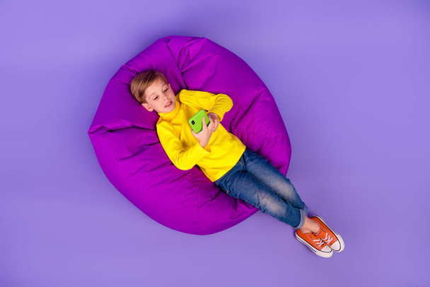 Full length photo top angle view of positive son wear yellow turtleneck sitting beanbag typing device isolated violet color background. - Photo, image