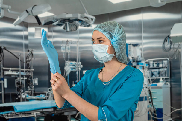 Bir kadın hemşire, karmaşık bir ameliyat sırasında kauçuk lateks mavi eldiven giyerken maske takan mavi bir üniforma giyer. İlaç. Doktorlar ameliyathane - Fotoğraf, Görsel