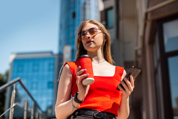 A business woman with a cup of coffee is talking on her smartphone while standing in the middle of the city center. The concept of a woman who earns money - 写真・画像