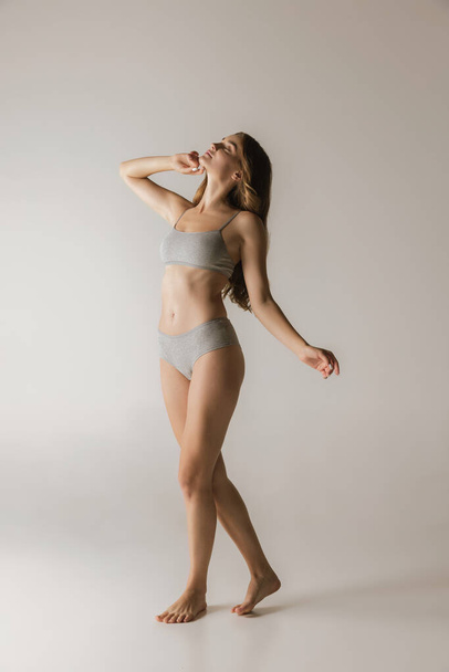 Slim sportive young woman in underwear on grey background. Fitness, diet, skin and body care. Fashion model with long curly hair. Copy space for ad - Φωτογραφία, εικόνα