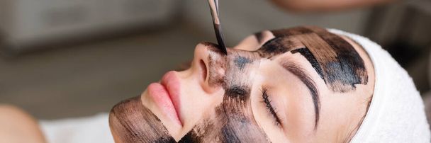 cosmetólogo aplicando máscara negra en cara de mujer bonita usando guantes negros, mujer hermosa en spa teniendo procedimientos faciales
.  - Foto, imagen