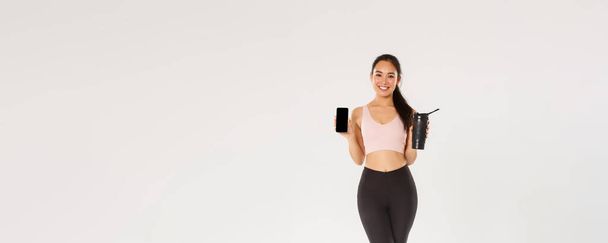 Πλήρες μήκος του χαμογελαστού όμορφη ασιατική fitness girl, αθλήτρια σε activewear δείχνει οθόνη smartphone και μπουκάλι νερό, τον έλεγχο πόσιμο υγρό με την εφαρμογή για κινητά κατά τη διάρκεια της προπόνησης. - Φωτογραφία, εικόνα