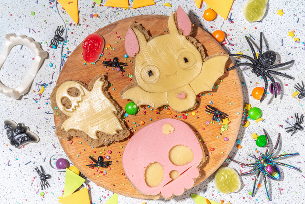Funny halloween sandwiches. Set of various decorated symbols sandwich - bat, skull, ghost, with cucumber, cheese and meat sausages, white table background with Halloween decorations - Fotó, kép