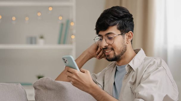 Close-up arabic hispanic guy young man wearing glasses bearded male user blogger sitting at home with mobile phone chatting online browsing web typing message smiling uses smartphone free wi-fi indoor - Zdjęcie, obraz