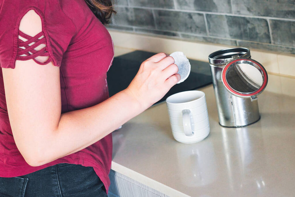 Uma jovem mulher adicionando um teabag redondo a uma caneca em uma cozinha doméstica moderna - Foto, Imagem