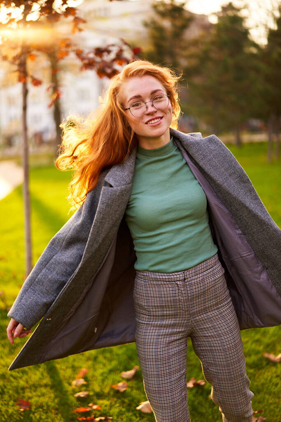Redhead smiling pretty woman walking in park on sunny autumn day. Elegant female poses in stylish authentic outfit has fun laughing on green lawn with vivid foliage, leaves. Girl enjoys a fall season. - Фото, изображение