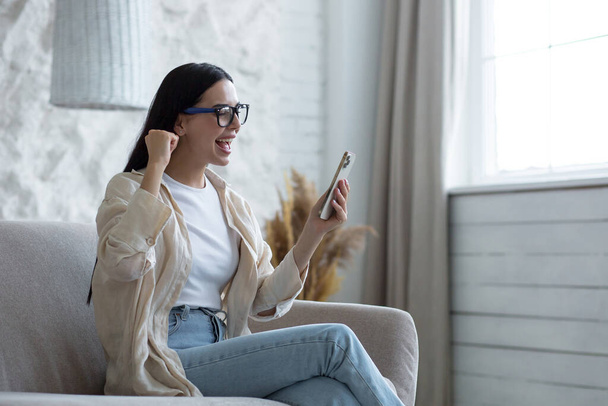 Happy young beautiful woman in glasses sitting on sofa at home with phone in hands. Celebrates a successful victory and winning the lottery, shouts wow, points with his hand yes. - Photo, Image