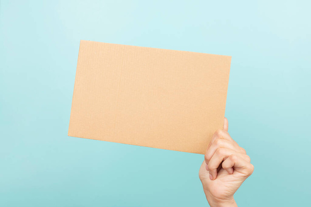 Woman hand holding blank sheet of brown cardboard on light blue background. - Photo, image