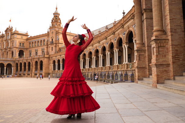 Beautiful teenage woman dancing flamenco in a square in Seville, Spain. She wears a red dress with ruffles and dances flamenco with a lot of art. Flamenco cultural heritage of humanity. - Foto, imagen