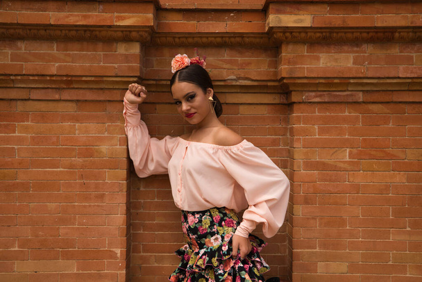 A beautiful teenage flamenco dancer dressed in a flowery skirt with ruffles and flowers in her hair is leaning against a red brick wall while making different body expressions. - Foto, immagini