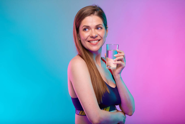 Smiling sporty woman in sportswear with water glass looking back over shoulder. Female fitness portrait isolated on neon multicolor background. - Φωτογραφία, εικόνα