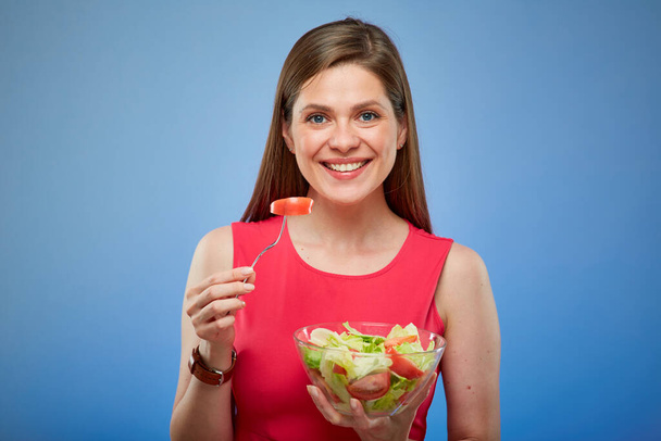 Young woman in red dress eating green salad with fork. Isolated female portrait on blue background. - Fotoğraf, Görsel