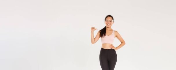 Volle Länge der selbstbewusst lächelnden asiatischen Athletin in Sport-BH und Leggins, Fitness-Girl zeigt auf sich selbst, führt einen aktiven und gesunden Lebensstil, ist professionell, steht weißer Hintergrund. - Foto, Bild