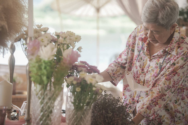 ηλικιωμένη γυναίκα διακοσμούν μπουκέτο λουλουδιών σε γυάλινο βάζο στο τραπέζι - Φωτογραφία, εικόνα