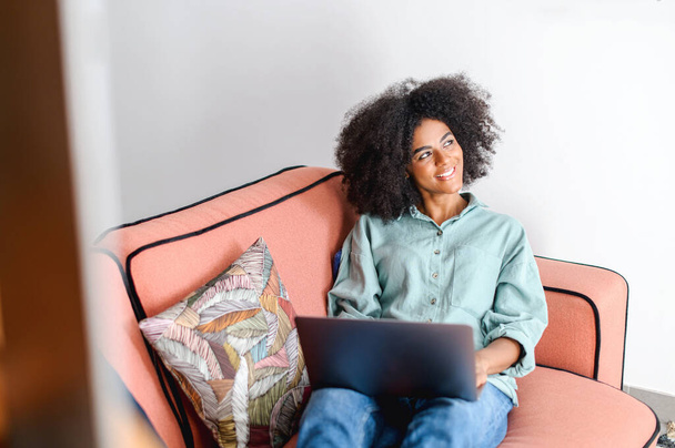 Optimistic african-american woman in casual wear using laptop while sitting on the couch. Young female student watching webinars, educational courses, learning on the distance, typing emails - Valokuva, kuva