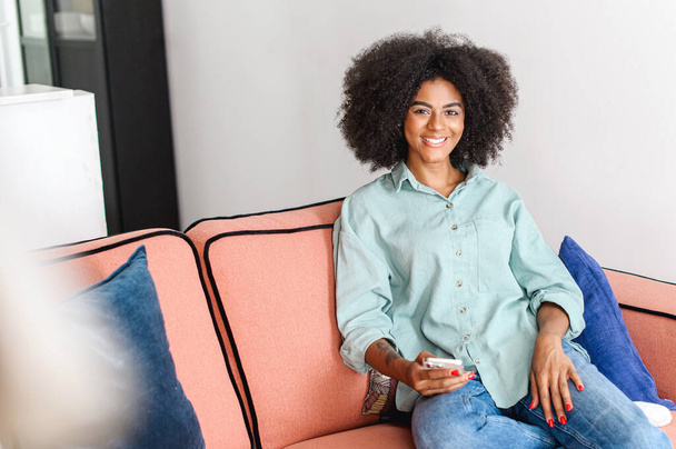 Portrait of beautiful black woman with natural curly afro hairstyle looking at the camera in cozy home atmosphere, charming serene and carefree girl holding smartphone and rest at home - Фото, зображення