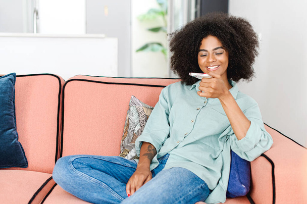 Young african woman sending voice message, recording note through smartphone sitting on the sofa in cozy modern apartment at home, using voice recognize feature - Foto, afbeelding