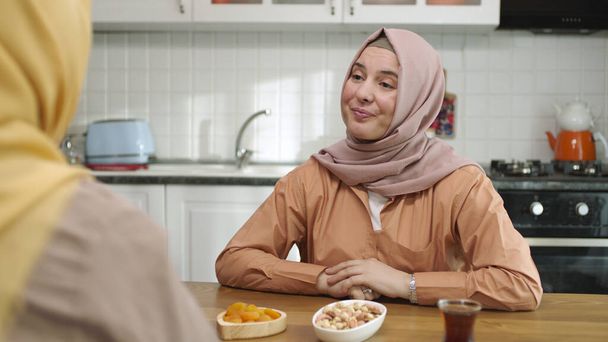 Blick über die Schulter einer muslimischen Hidschab-bekleideten Frau, die sich am Küchentisch mit ihrem Freund unterhält. Porträt zweier junger Freundinnen, die sinnvolle Gespräche in der Küche führen. - Foto, Bild