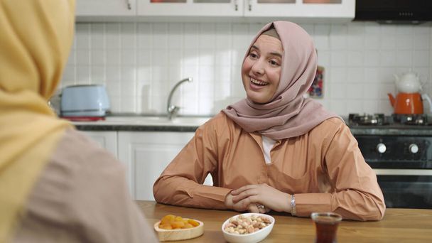 Vue au-dessus de l'épaule d'une musulmane habillée en hijab discutant avec son ami à la table de la cuisine. Portrait de deux jeunes amies ayant des conversations significatives dans la cuisine. - Photo, image